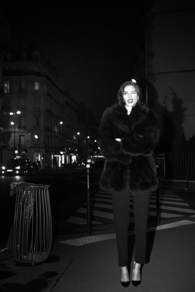 Cette mannequin se tient debout avec assurance dans une rue de Paris faiblement éclairée la nuit, vêtue d'un manteau de fourrure sombre et luxueux et d'un pantalon noir. L'arrière-plan urbain présente des immeubles et des lampadaires doucement éclairés, créant une atmosphère maussade.