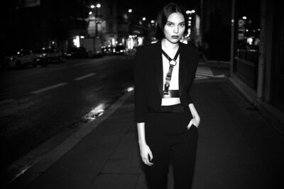 Cette mannequin vêtue d'une élégante tenue noire marche avec assurance dans une rue déserte de Paris, la nuit. La rue est faiblement éclairée par des lampadaires lumineux et il y a des immeubles en arrière-plan. La scène dégage une atmosphère urbaine sophistiquée.