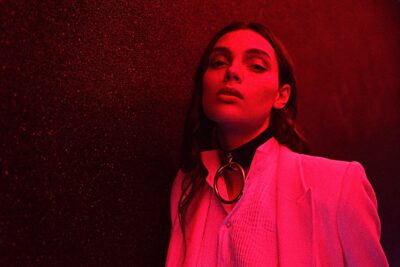 Cette mannequin en blazer rose se tient debout devant un mur sombre, éclairé par une lumière rouge. Elle regarde la caméra avec assurance, vêtue d'un chemisier blanc et d'un collier ras du cou avec une grosse bague en argent.
