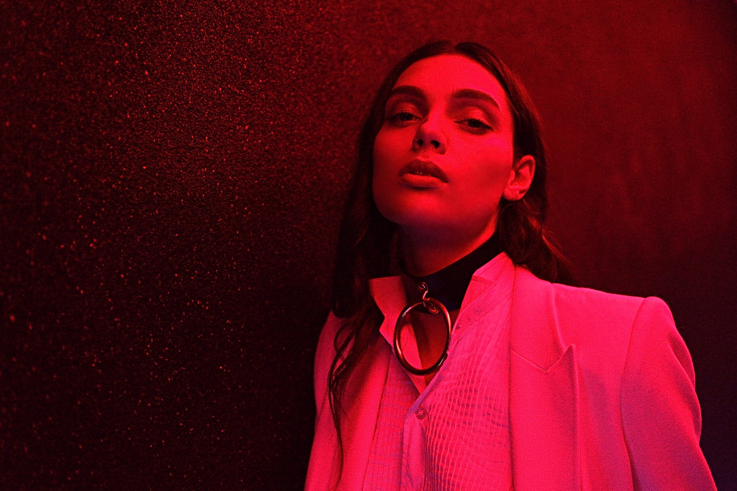 Cette mannequin en blazer rose se tient debout devant un mur sombre, éclairé par une lumière rouge. Elle regarde la caméra avec assurance, vêtue d'un chemisier blanc et d'un collier ras du cou avec une grosse bague en argent.