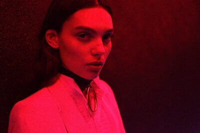 Cette mannequin aux cheveux longs, portant un blazer blanc et un collier ras du cou, se tient contre un mur sombre baigné de lumière rouge, regardant directement la caméra.