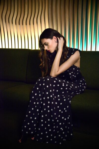 Cette mannequin assise sur un canapé vert foncé, vêtue d'une longue robe noire avec des étoiles blanches. Elle penche la tête vers le bas, sa main posée sur son cou, dans une pièce doucement éclairée avec un mur en bois texturé en arrière-plan.