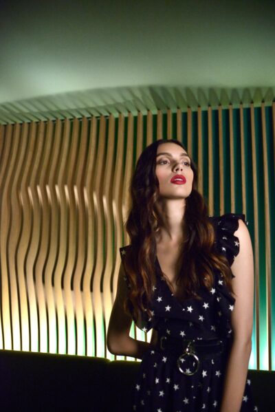 Cette mannequin aux cheveux longs et portant une robe à motifs étoilés se tient devant un mur ondulé en bois lambrissé avec un éclairage vert.